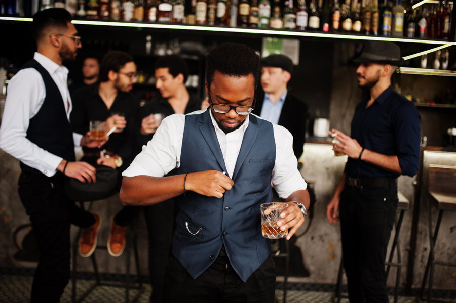 A man in glasses buttons his vest, with a drink in his hand, in a pub.