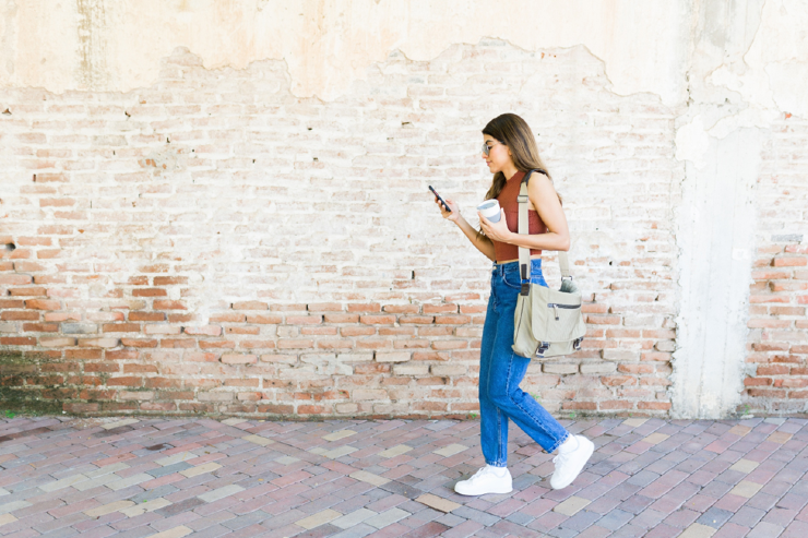 A woman walking near a brick wall, cell phone and coffee cup in hand.