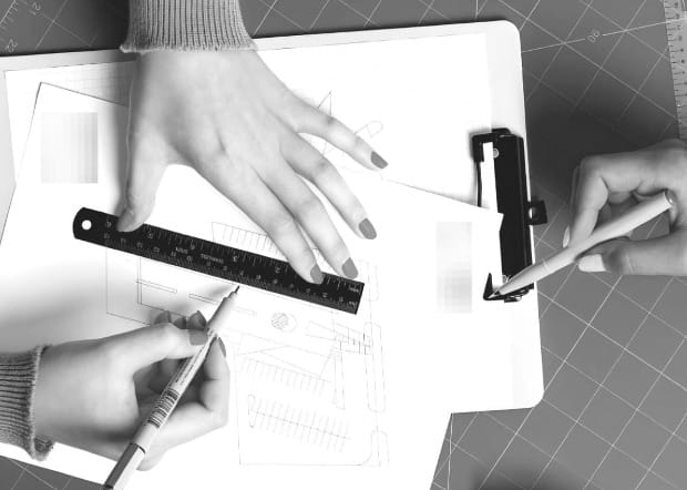two sets of hands work on engineering documents, one has a ruler in hand, the other is holding a pen.