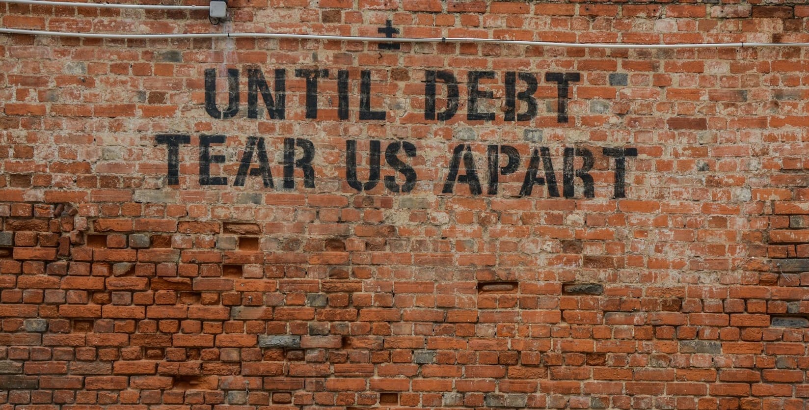 “Graffiti on a brick wall reads ‘UNIT DEBT TEAR US APART.’”