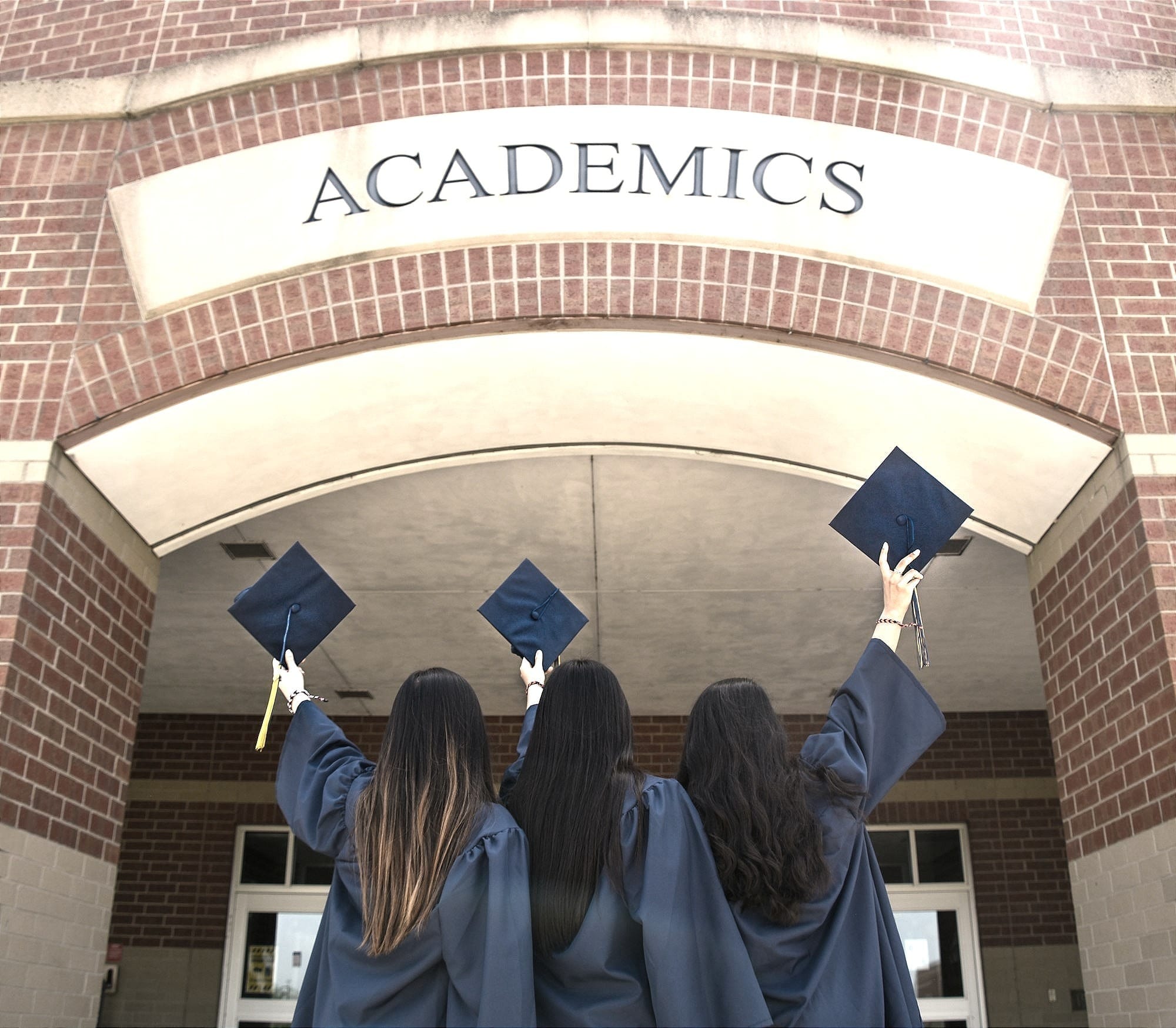 Three graduates in caps and gowns raise their mortarboards beneath a brick arch labeled ‘ACADEMICS.’