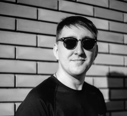 A man in black crew neck shirt wearing black sunglasses and smiling slightly, thinking cha-ching.