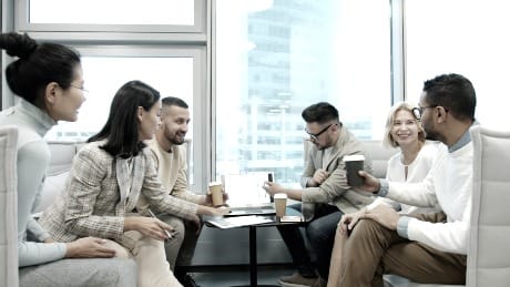 A small group of colleagues sit in a lounge area talking over coffee, with a city view through large windows.