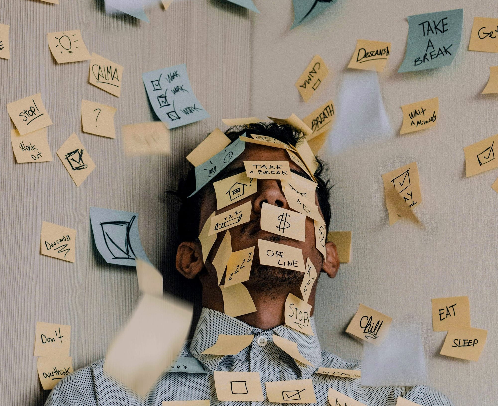A man leaning against a wall covered in sticky notes with reminders and commands, several notes stuck to his face, conveying information overload and mental clutter.