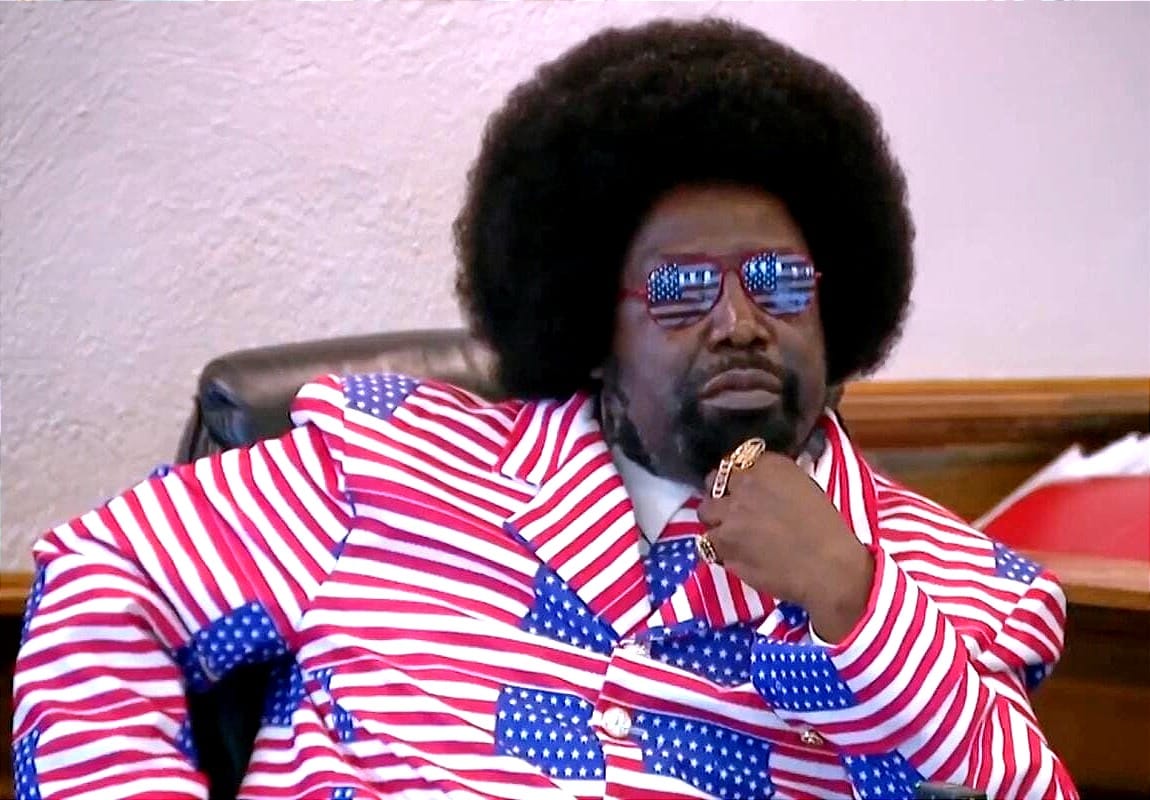 Afroman Joseph Foreman seated in court wearing a red, white, and blue American flag suit and matching flag-pattern sunglasses, resting his hand near his chin.