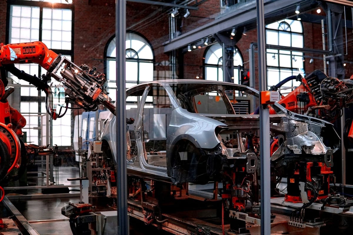 Robotic arms operate on a car body frame inside an industrial manufacturing facility with large windows.