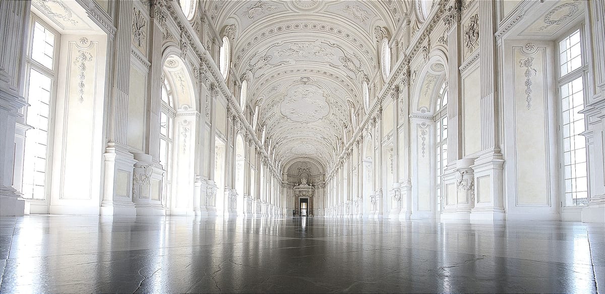 A long, ornate, sunlit hall with high arched ceilings, tall windows, and a polished floor leading toward a distant doorway.
