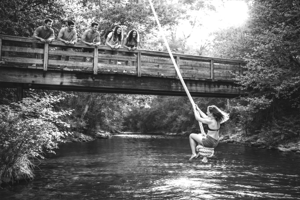 A teenage girl sitting on a rope swing at the high point of its arc above a river, watched by friends standing on a wooden bridge on a summer day.