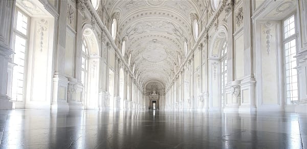 A long, ornate, sunlit hall with high arched ceilings, tall windows, and a polished floor leading toward a distant doorway.