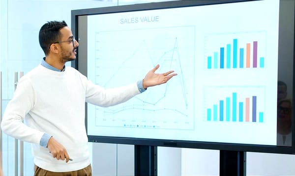 A person gestures toward a large screen displaying charts and bar graphs labeled “Sales Value.”